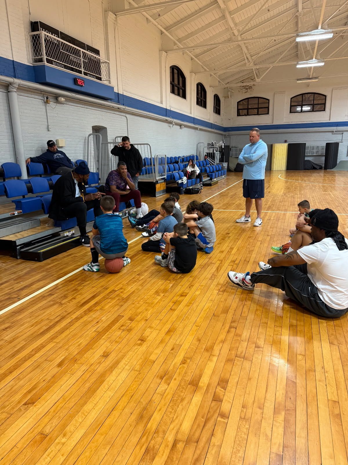Kids sitting on the floor of an old school gym while Coach Ray talks
